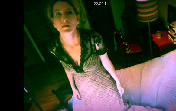 Heathen Derr (Heather Derr-Smith) Film Still. The poet is standing on a bed looking up into a surveillance camera that is watching them. The poet is wearing a ripped lace dress, arms to the side, face in a state of dread or drugged-up expression.