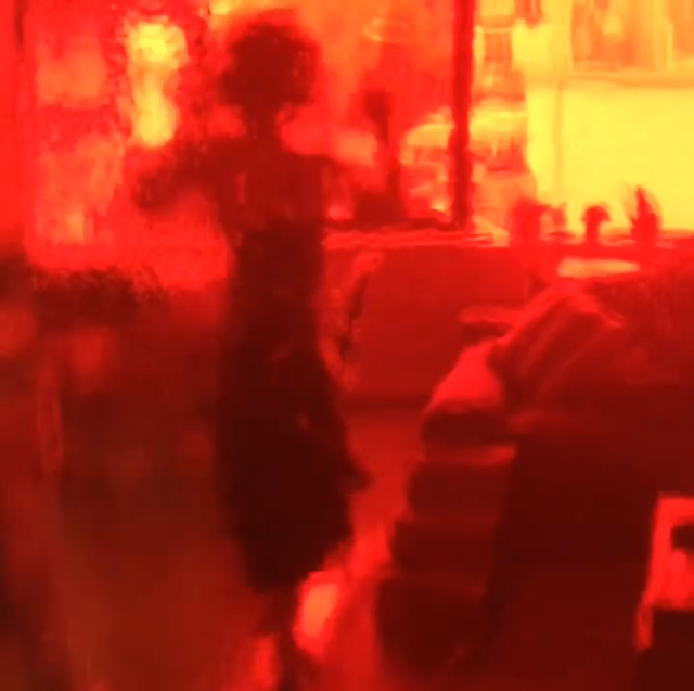 Image is of a film still of Heathen Derr (Heather Derr-Smith) dancing along with the Madison Dance Scene in Bande a Parte by Godard. The camera is filming through a red bottle so everything is tinted red and distorted by the glass.