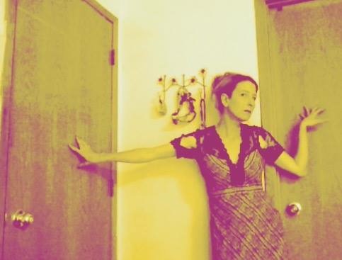 Film still of Heathen Derr (Heather Derr-Smith) The poet is standing  with their back and outstretched arms against two doors with their right arm bent, palm pressed behind them against a door to the right and the left arm reaching out to press the door to the left. The poet is wearing a ripped lace dress. There is the jaw bone of a shark hanging behind them to the left.