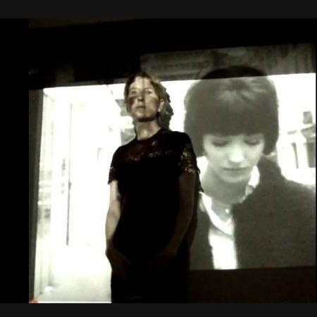 Heathen Derr (Heather Derr-Smith) film still. The poet stands in front of a movie screen upon which the film Vivre sa Vie is being projected. The film is projected also on the poet's body. You can see Anna Karina on the screen behind the poet. The poets face is partially obscured by the projected image but they stare out at something beyond the frame.